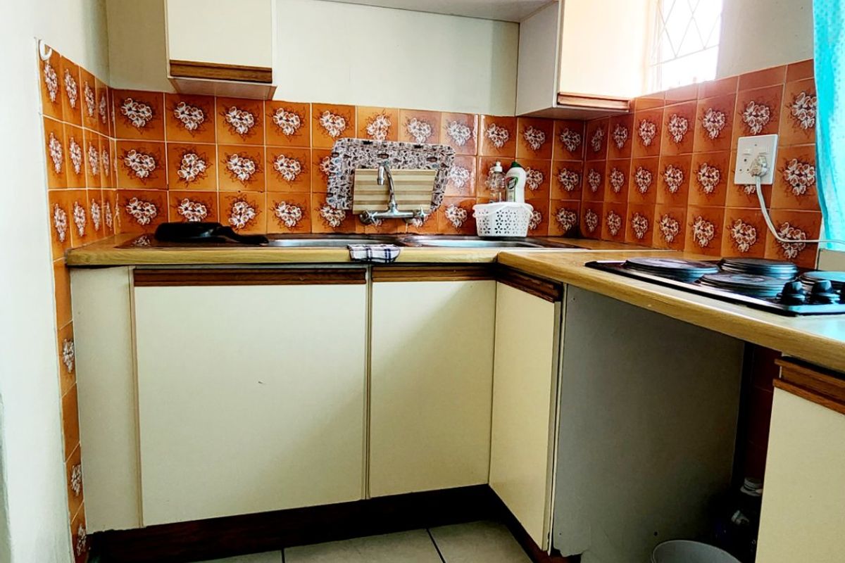 A compact kitchen featuring dated cabinetry, orange patterned tile backsplash, ceramic floor tiles, and a built-in hob with integrated sink area.