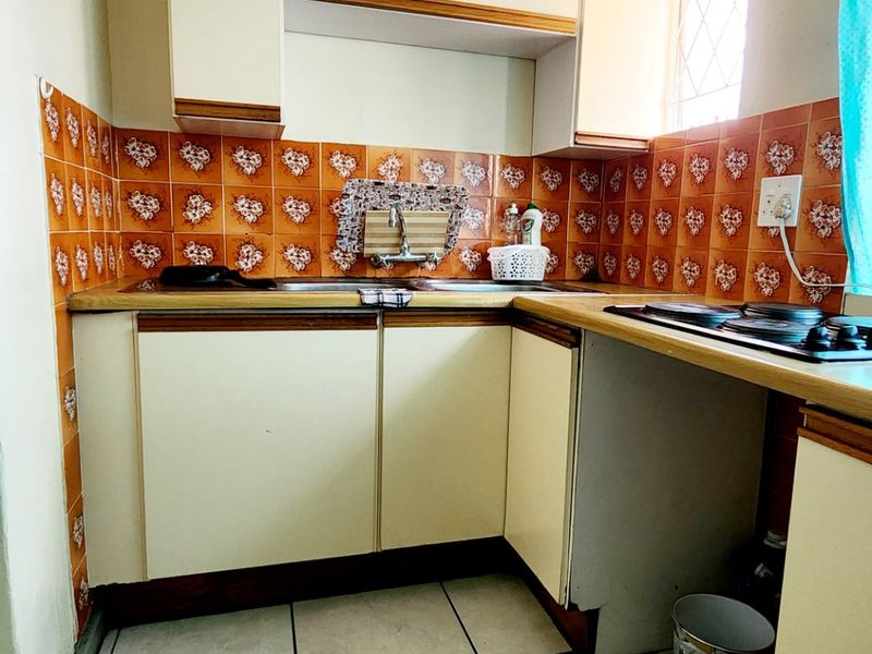 A compact kitchen featuring dated cabinetry, orange patterned tile backsplash, ceramic floor tiles, and a built-in hob with integrated sink area.