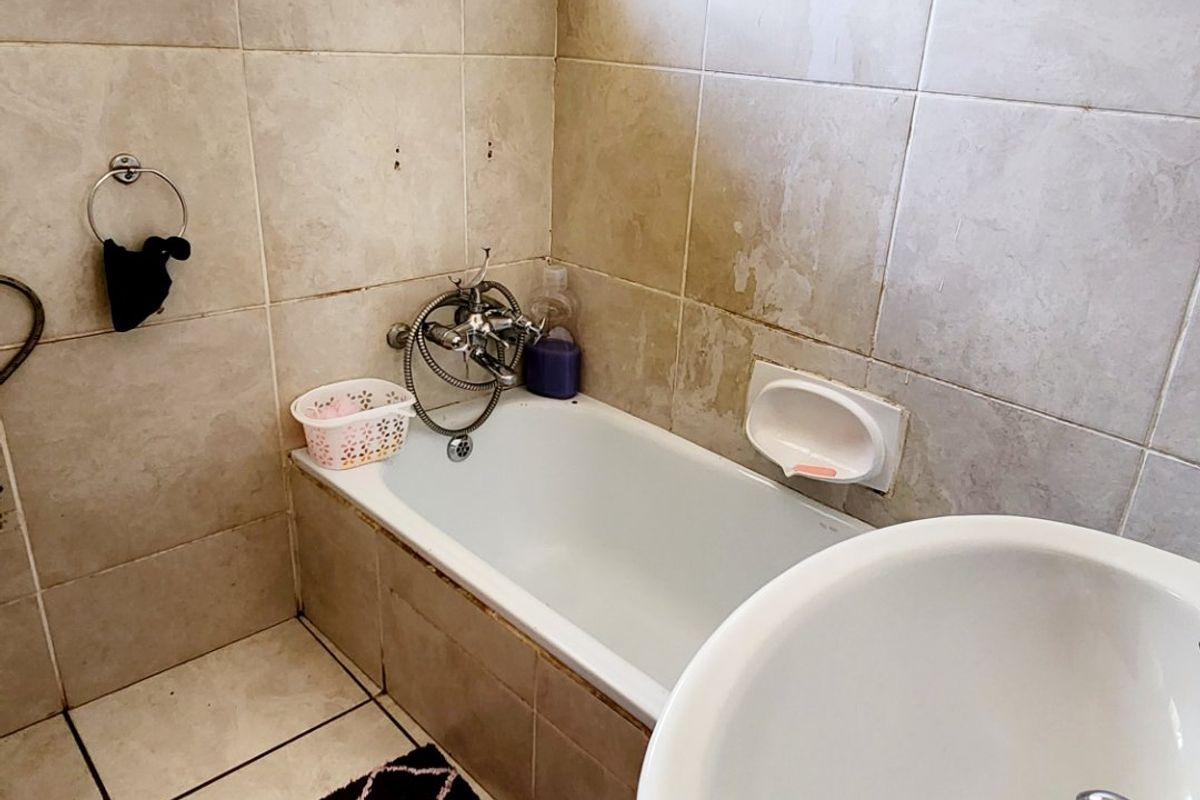 A compact bathroom featuring a bathtub, pedestal basin, and neutral ceramic tiling. The space requires cosmetic updates to finishes and grout.