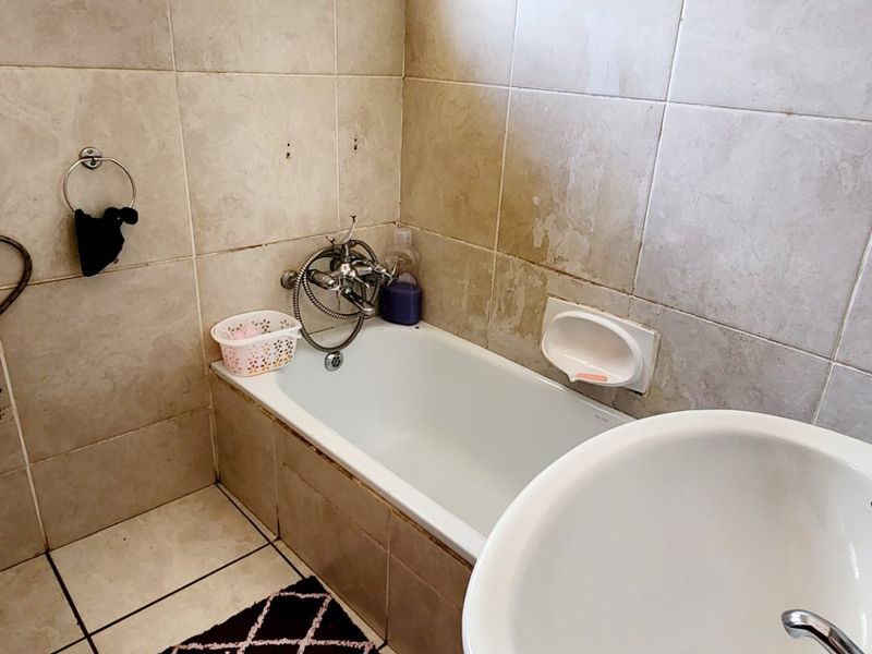 A compact bathroom featuring a bathtub, pedestal basin, and neutral ceramic tiling. The space requires cosmetic updates to finishes and grout.