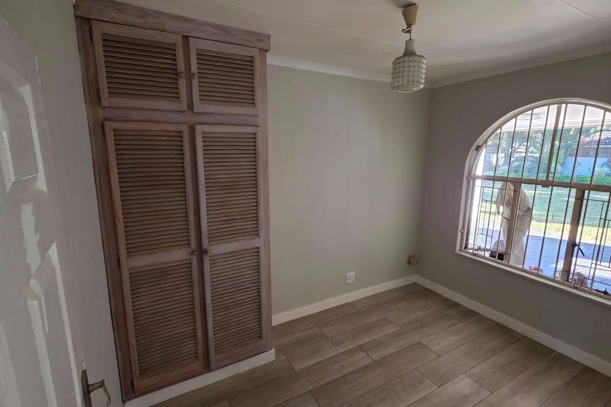 A bright, empty bedroom featuring wood-look tile flooring, a built-in wardrobe with louvered doors, and a distinctive arched window with security bars.