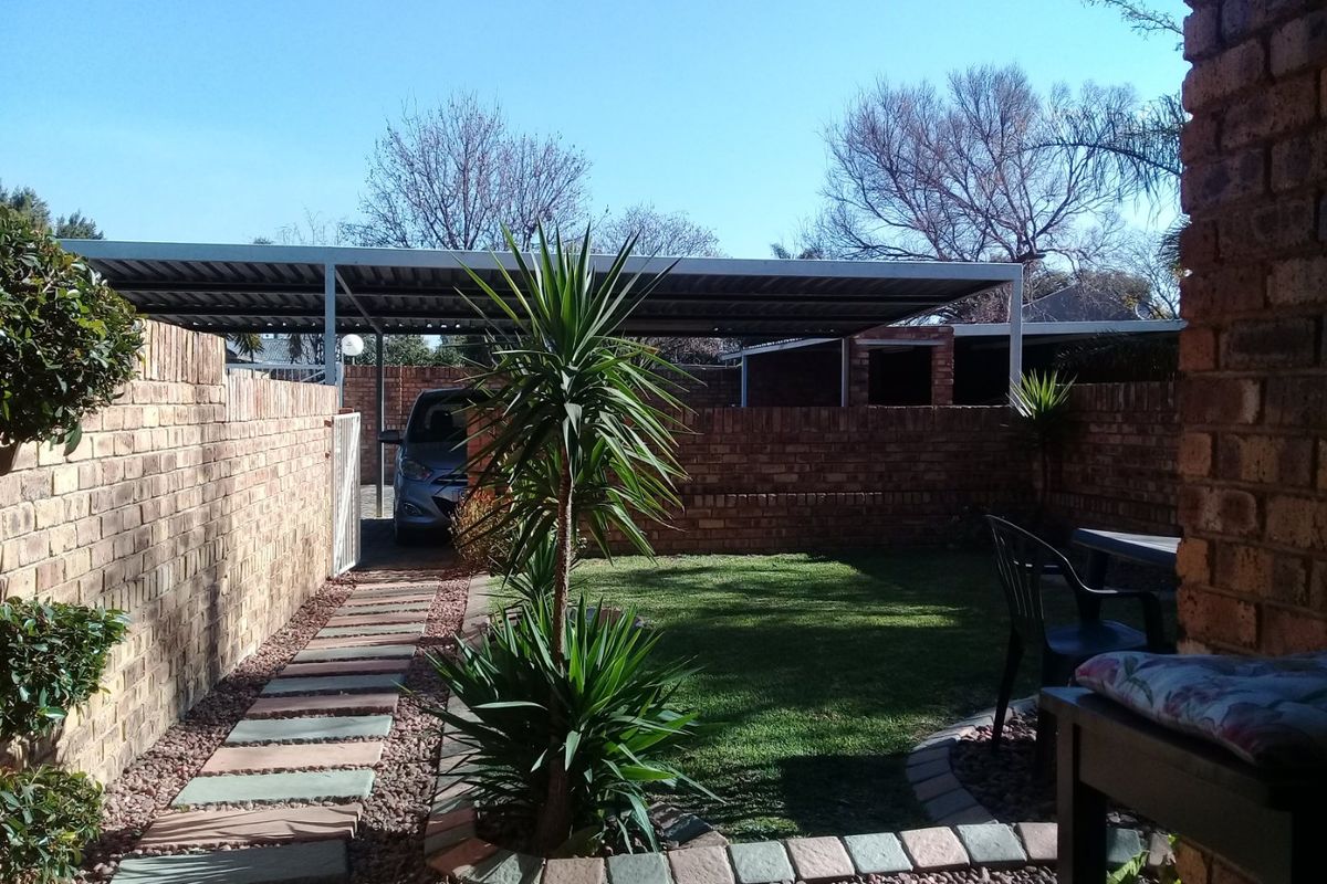 A private rear garden featuring a brick boundary wall, a paved walkway, a small lawn area, and a covered carport structure.