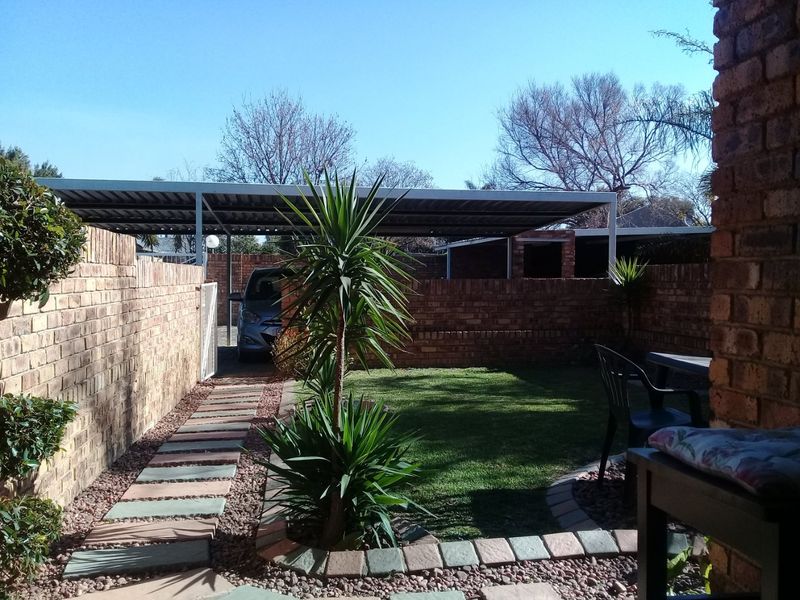 A private rear garden featuring a brick boundary wall, a paved walkway, a small lawn area, and a covered carport structure.