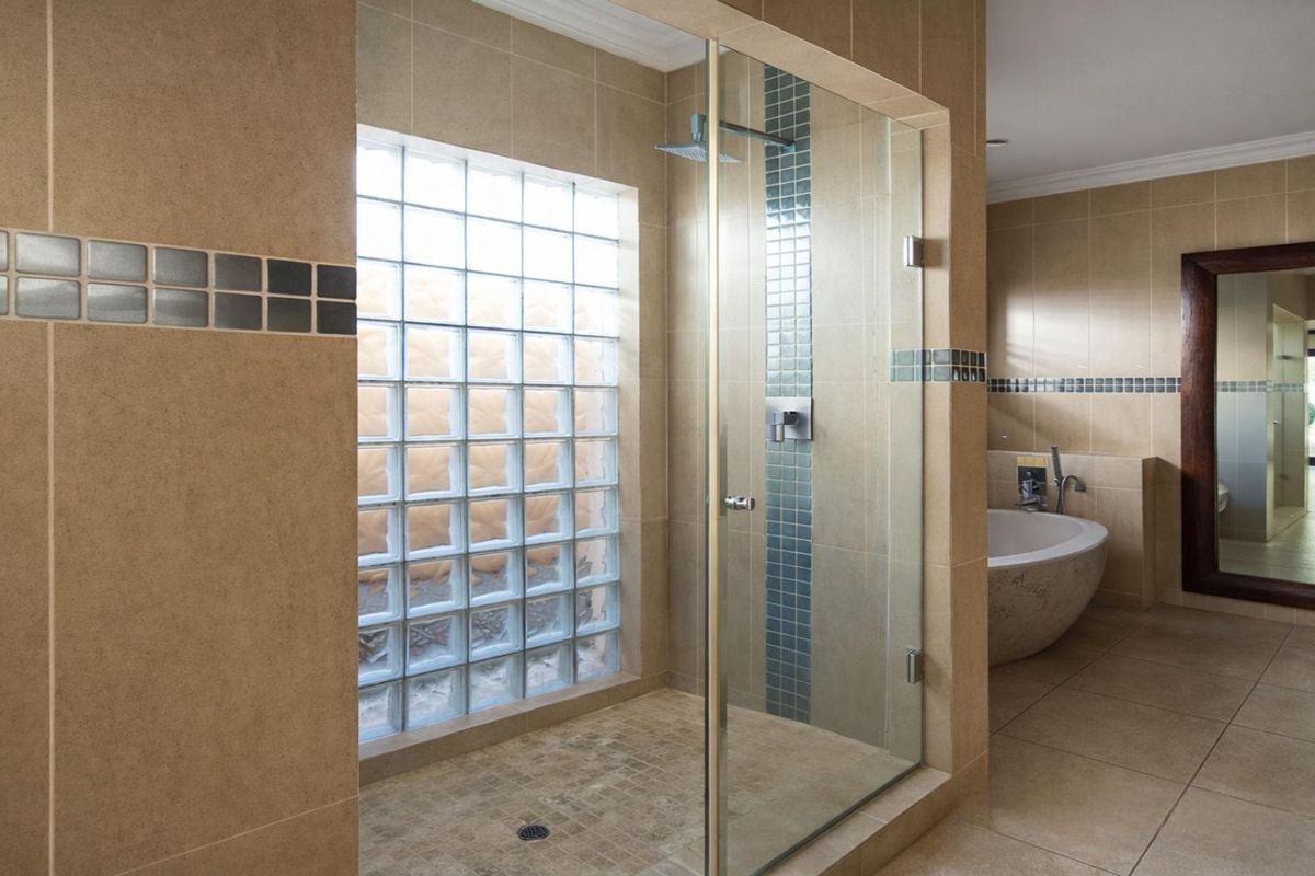 Spacious bathroom featuring a glass-enclosed shower with a glass block window, a freestanding bathtub, and neutral wall and floor tiling.