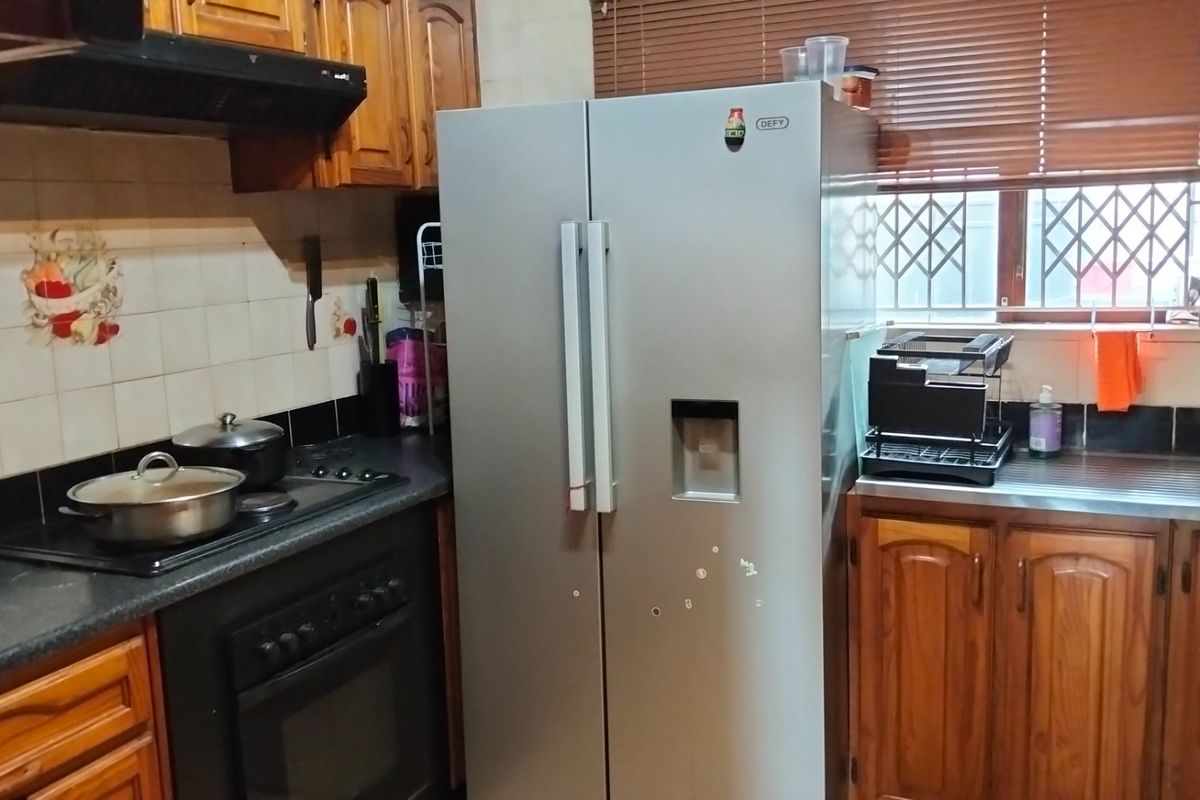 A functional kitchen featuring wooden cabinetry, laminate countertops, built-in oven, hob, extractor fan, and a stainless steel sink with window security bars.