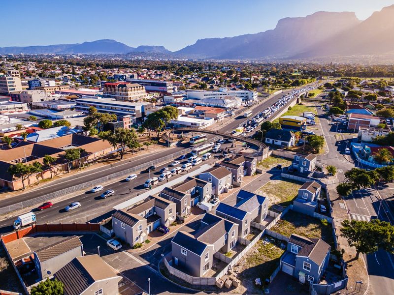 An aerial view of a residential housing development situated near a major road and urban infrastructure with mountain views in the background.