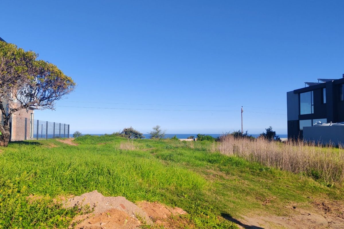 A vacant residential plot featuring natural grass coverage and distant ocean views, situated between existing modern residential properties.