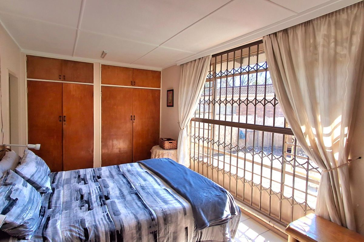 A bedroom featuring built-in wooden cupboards, tiled flooring, and a large window secured with a metal security gate.