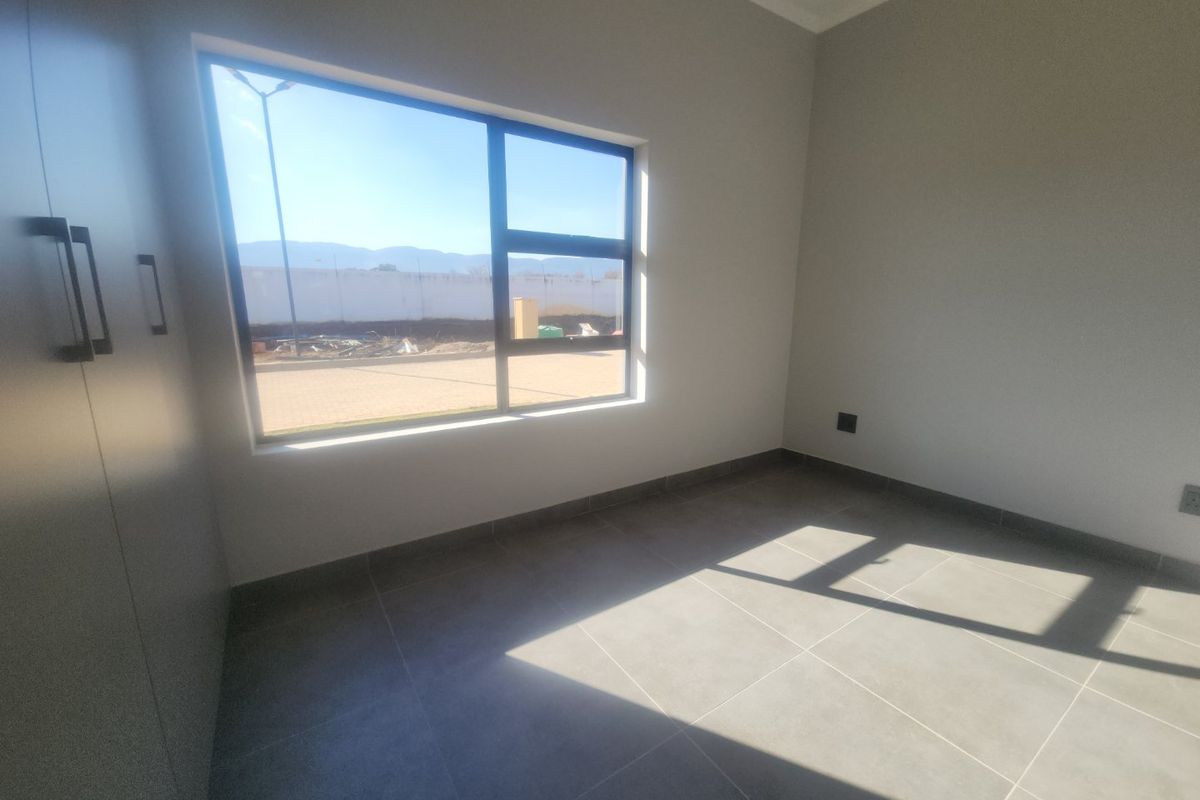 A bright, empty bedroom featuring modern grey floor tiles, built-in cupboards, and large aluminium-framed windows allowing for ample natural light.