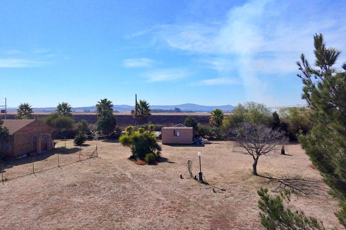 An aerial view of a large, dry residential plot featuring a detached outbuilding, perimeter fencing, and mature trees against a mountain backdrop.