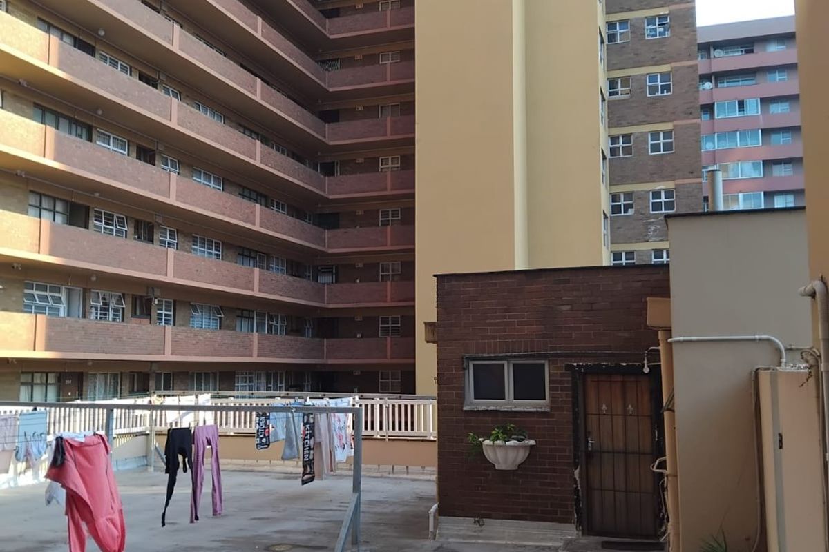 A rear exterior patio area featuring a concrete surface, brick wall detailing, and a security gate, overlooking a large apartment complex.