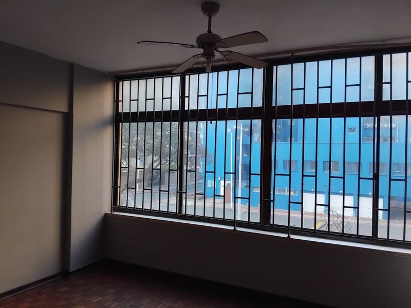 An empty living area featuring parquet flooring, a ceiling fan, and large windows fitted with security bars.