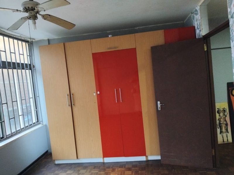 A bedroom featuring built-in cupboards with a red accent panel, parquet flooring, a ceiling fan, and security bars on the window.