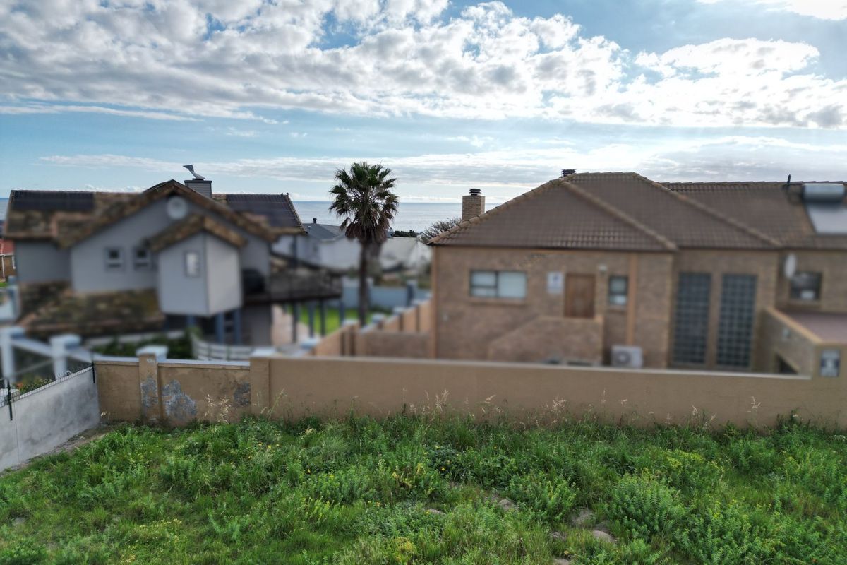 A view of a vacant residential plot featuring a boundary wall with visible wear, overlooking neighboring properties and a distant ocean view.