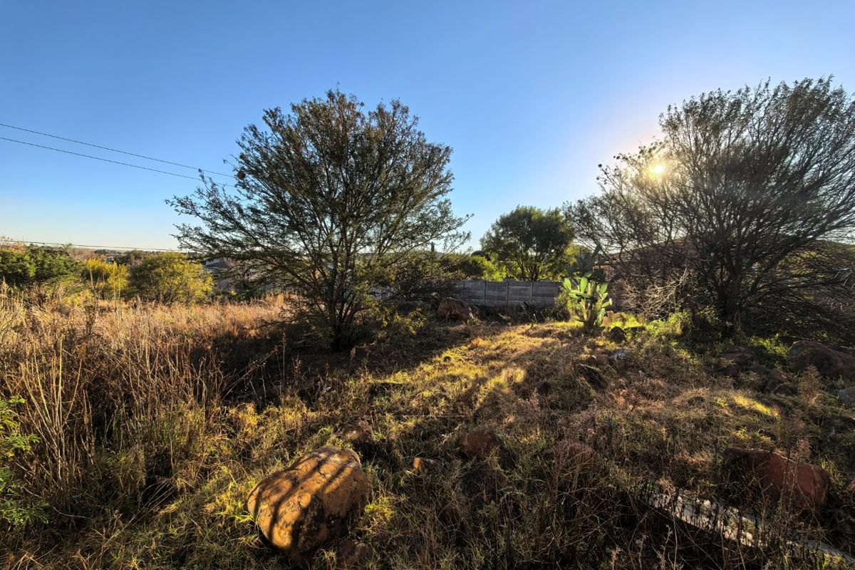A view of a vacant residential plot featuring natural rocky terrain, indigenous trees, and a precast concrete boundary wall.