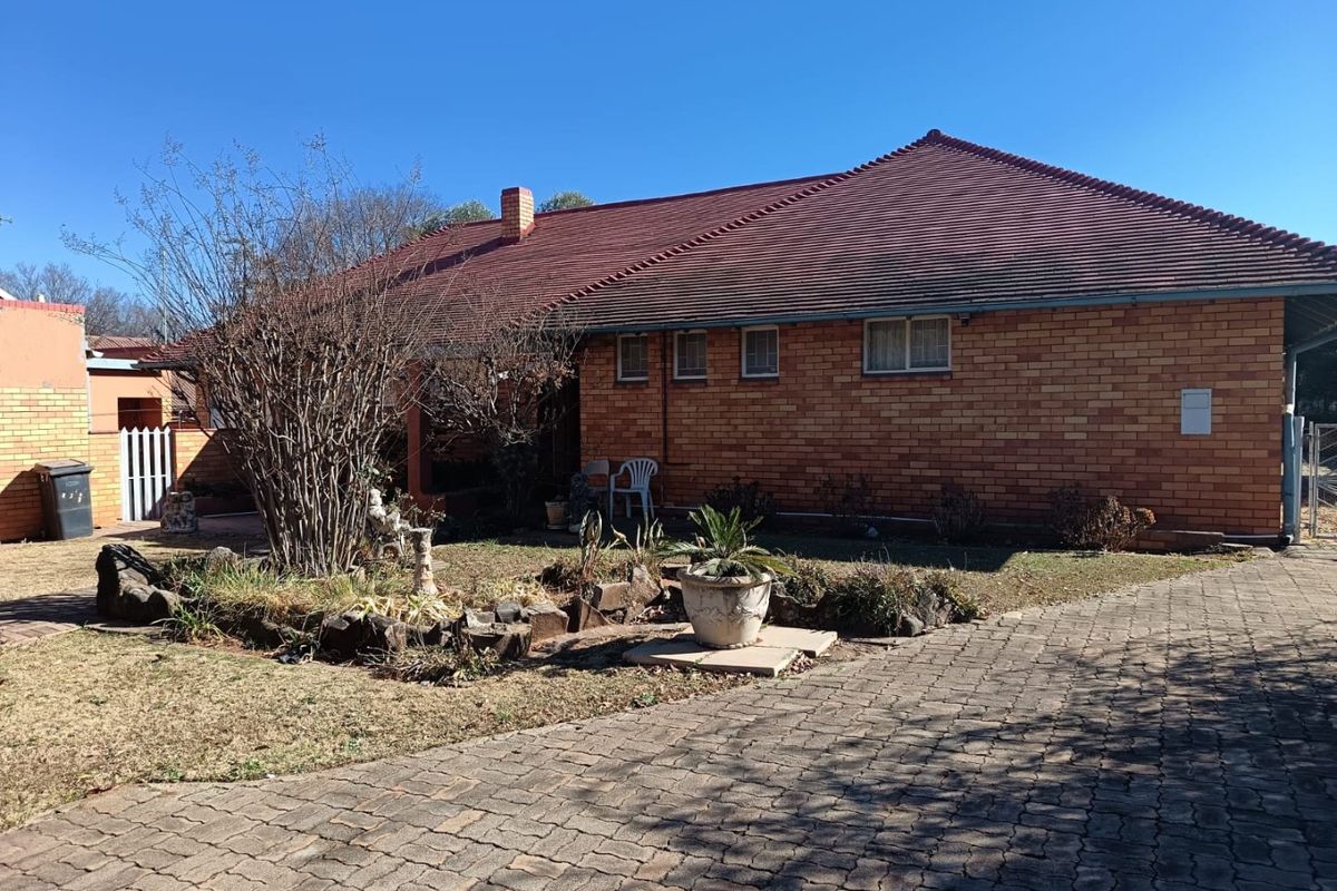 Rear view of a single-story brick house featuring a tiled roof, chimney, and a paved driveway leading to the garden area.