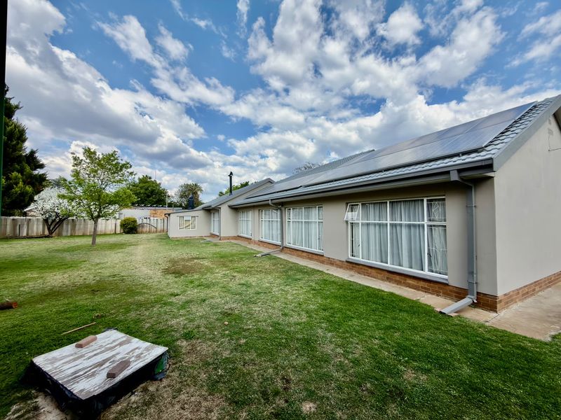 Rear view of the house featuring a large lawn, solar panel installation on the roof, and multiple windows along the exterior wall.