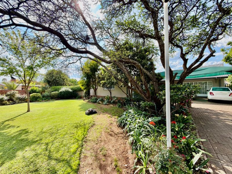A well-maintained front garden featuring a lush lawn, mature trees, and a paved driveway leading to the property entrance.