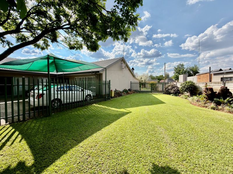 A well-maintained rear garden featuring a manicured lawn, perimeter fencing, and a shade sail covering a paved driveway area.