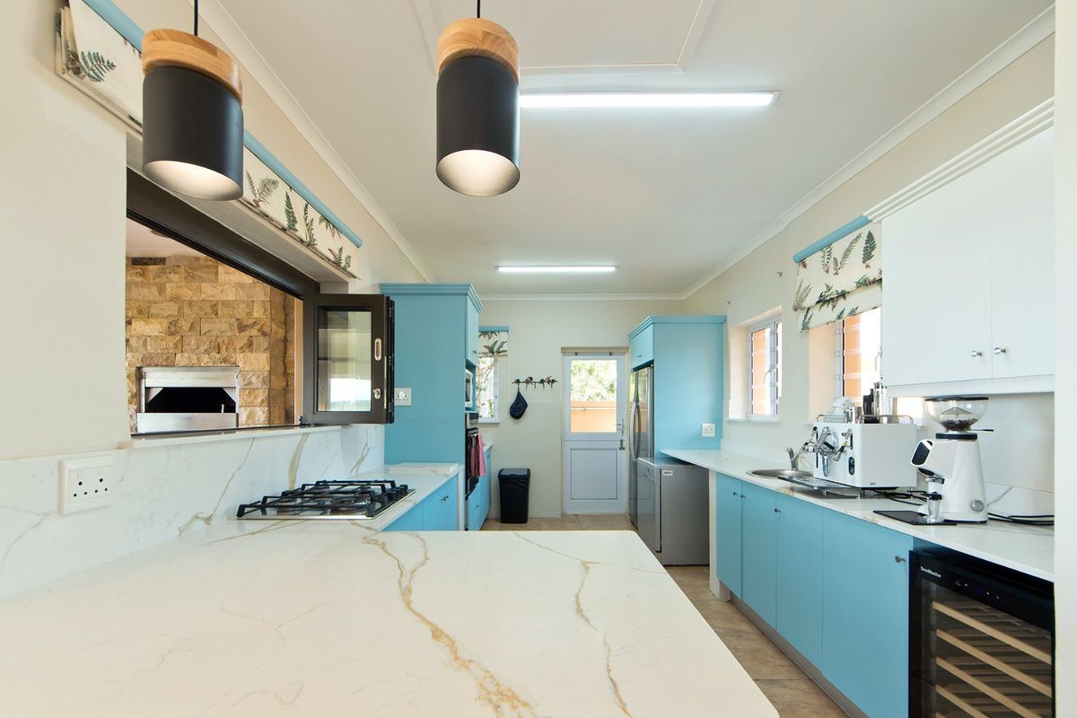 A bright, modern kitchen featuring light blue cabinetry, stone countertops, a gas hob, and a convenient pass-through window overlooking an outdoor area.