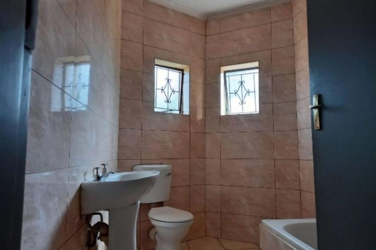 A dated bathroom featuring pink ceramic wall tiles, a pedestal basin, toilet, and bathtub with security bars on the windows.