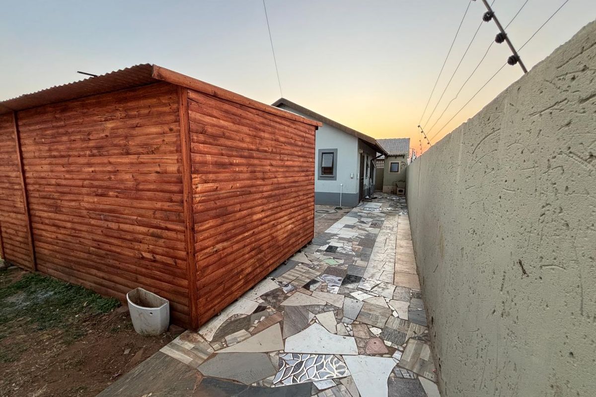 A side exterior view of the property featuring a wooden storage shed, a paved walkway, and a boundary wall with electric fencing.