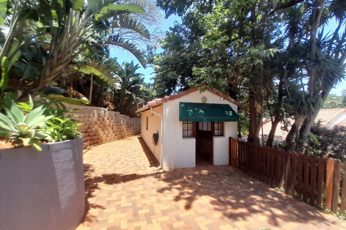 A well-maintained exterior view of a small outbuilding featuring a tiled roof, green awning, and paved access path surrounded by mature trees.
