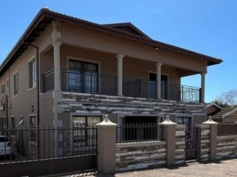 A two-story residential building featuring stone cladding, a spacious upper balcony with wrought iron railings, and a secure gated entrance with paved driveway.