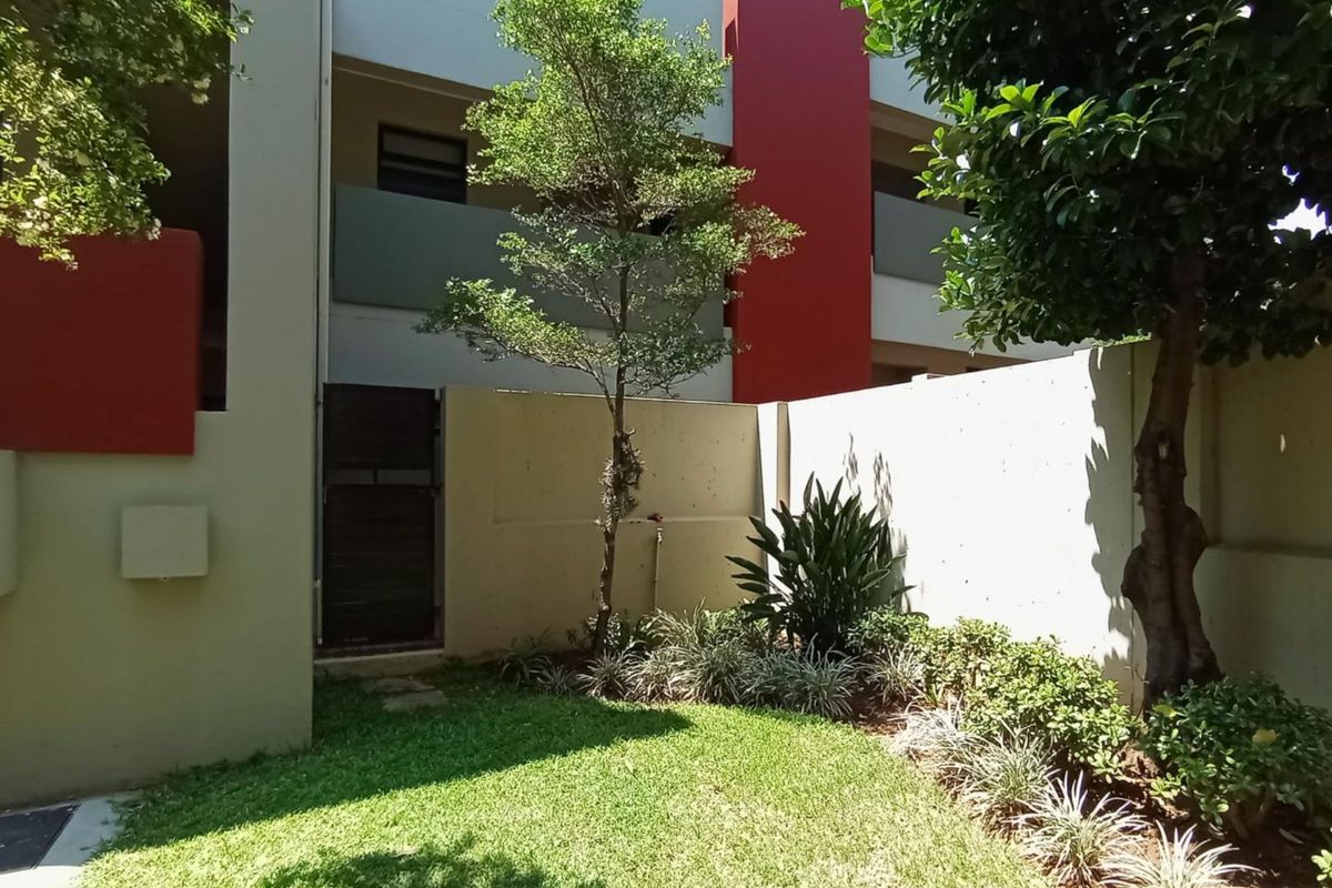 A private rear garden area featuring a manicured lawn, established trees, and a secure boundary wall within an apartment complex.