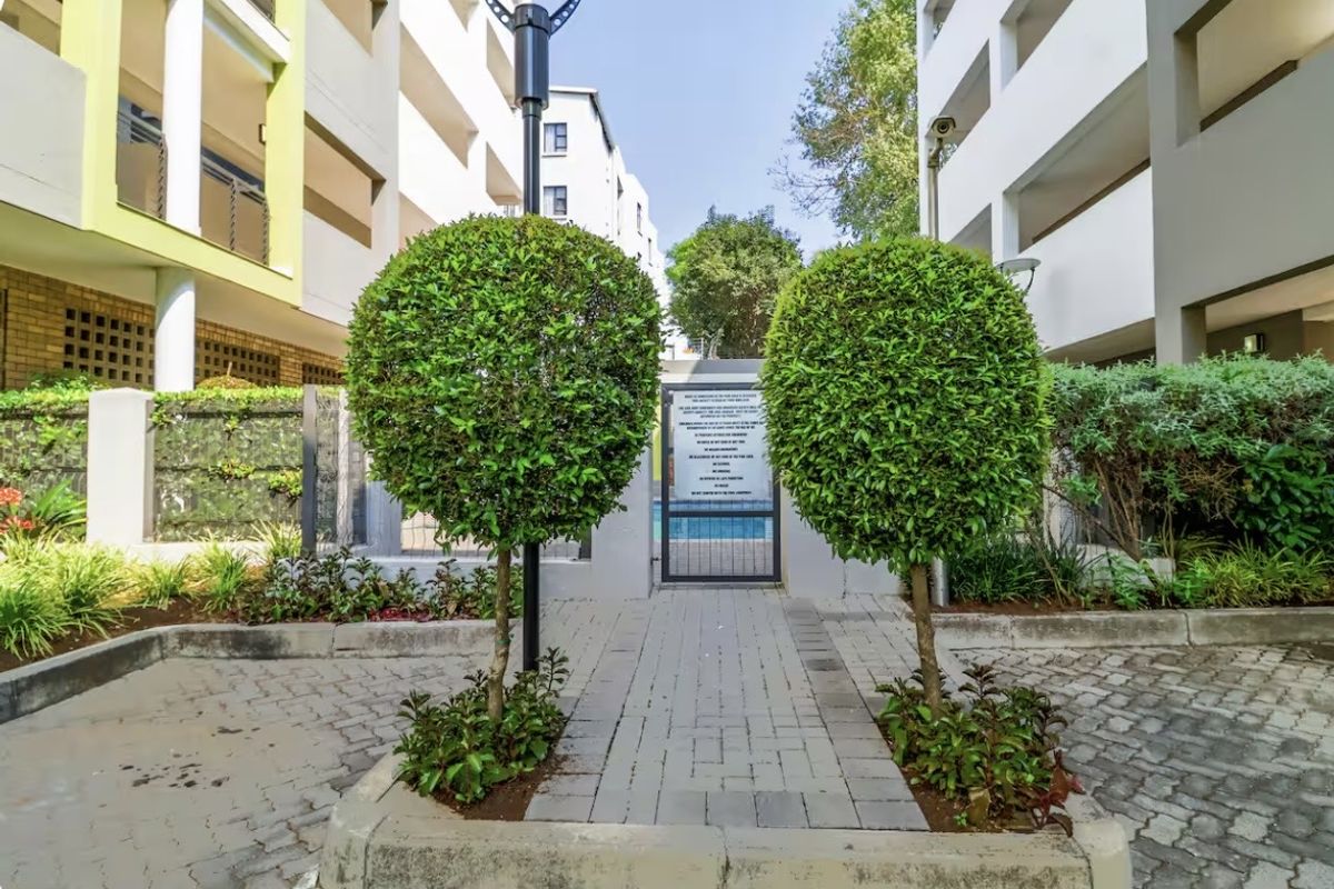 A well-maintained paved walkway leading through a landscaped garden area between apartment blocks, featuring a secure gate and exterior lighting.