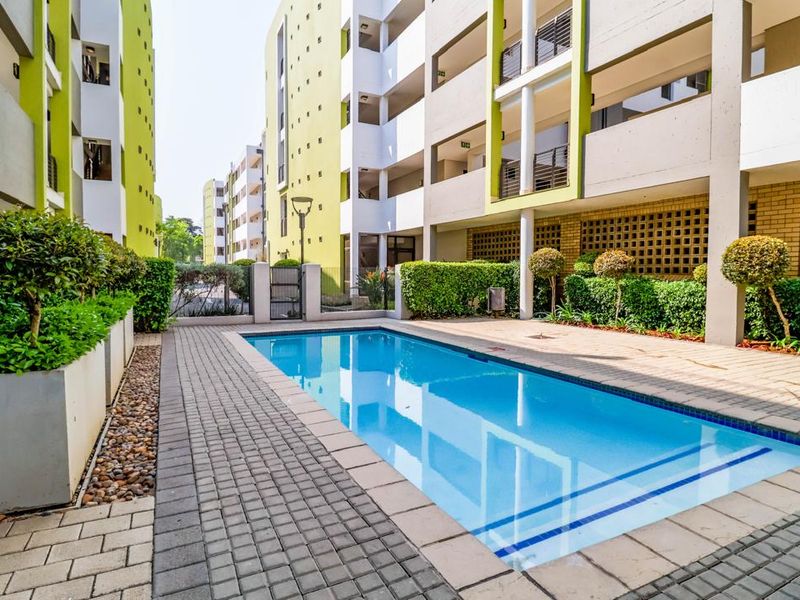 A well-maintained communal swimming pool area situated between modern apartment blocks, featuring paved decking and landscaped garden borders.
