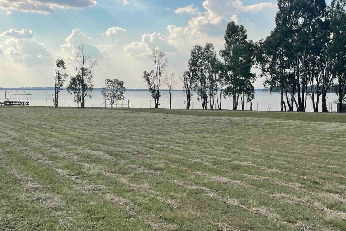 A scenic view of a large, open grassy plot overlooking a tranquil lake, framed by mature trees and perimeter fencing.