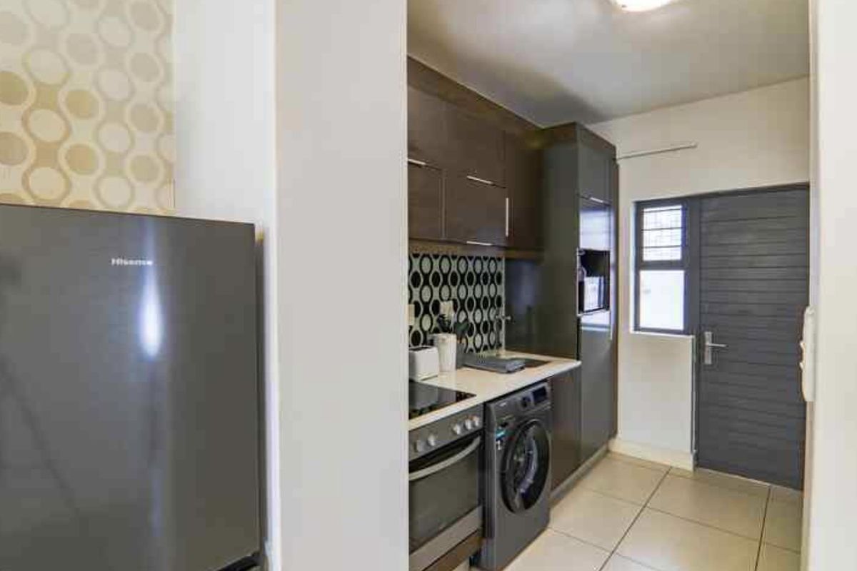 Compact modern kitchen featuring dark cabinetry, patterned backsplash, built-in oven, hob, and integrated laundry appliance with tiled flooring.