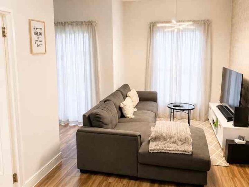 A bright and well-maintained living room featuring wood-look laminate flooring, modern pendant lighting, and large windows with neutral curtains.