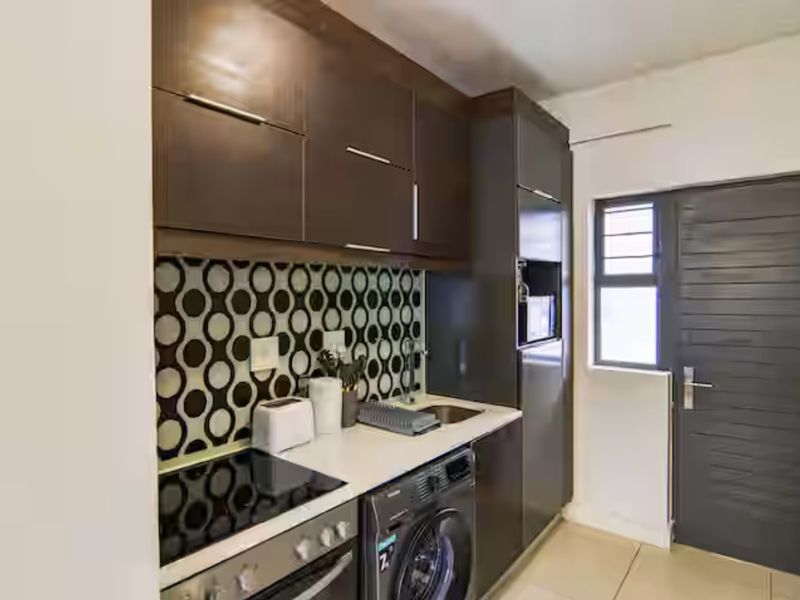 Compact kitchen featuring dark wood cabinetry, a patterned tile backsplash, integrated electric hob, oven, and tiled flooring.