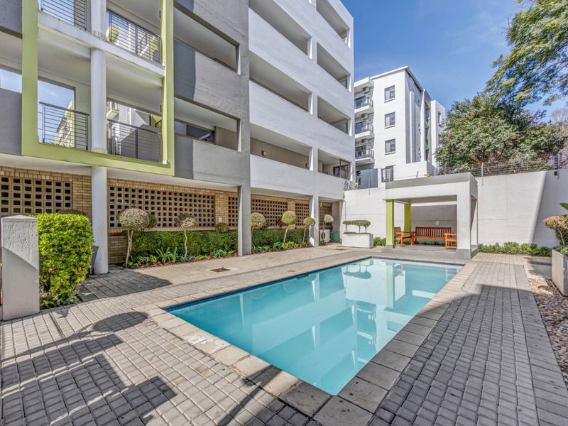 A well-maintained communal swimming pool area featuring paved decking, a covered seating structure, and landscaped garden beds set against the apartment building facade.