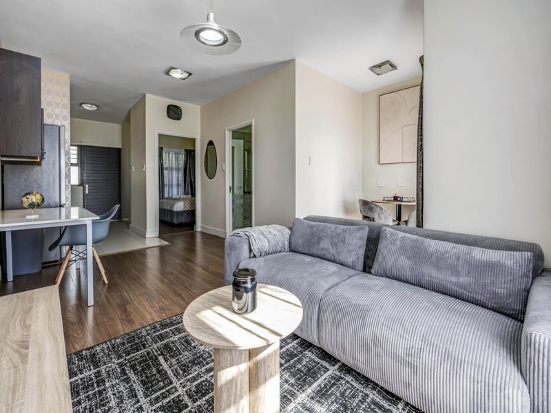 An open-plan living area featuring laminate flooring, recessed lighting, and a compact kitchen space with modern cabinetry, leading to adjacent rooms.