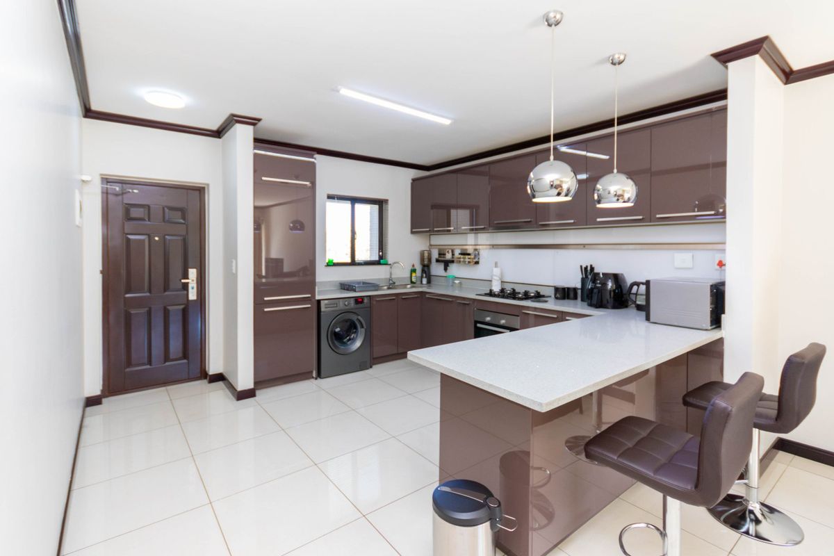 Modern kitchen featuring glossy brown cabinetry, stone countertops, a breakfast bar, built-in oven, gas hob, and tiled flooring in an open-plan layout.