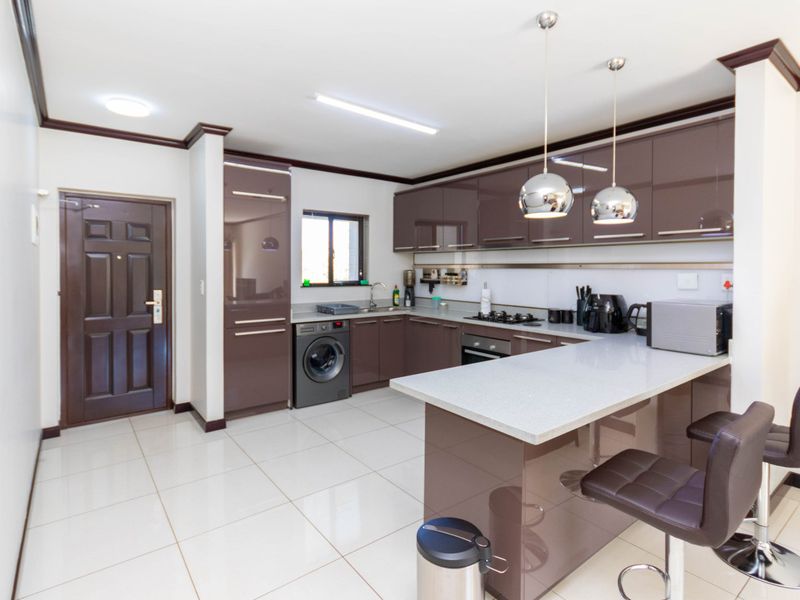 Modern kitchen featuring glossy brown cabinetry, stone countertops, a breakfast bar, built-in oven, gas hob, and tiled flooring in an open-plan layout.