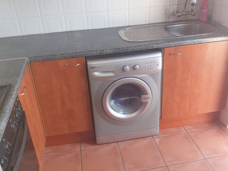 A functional kitchen area featuring wood-look cabinetry, granite-effect countertops, a stainless steel sink, and tiled flooring with space for a front-loading washing machine.