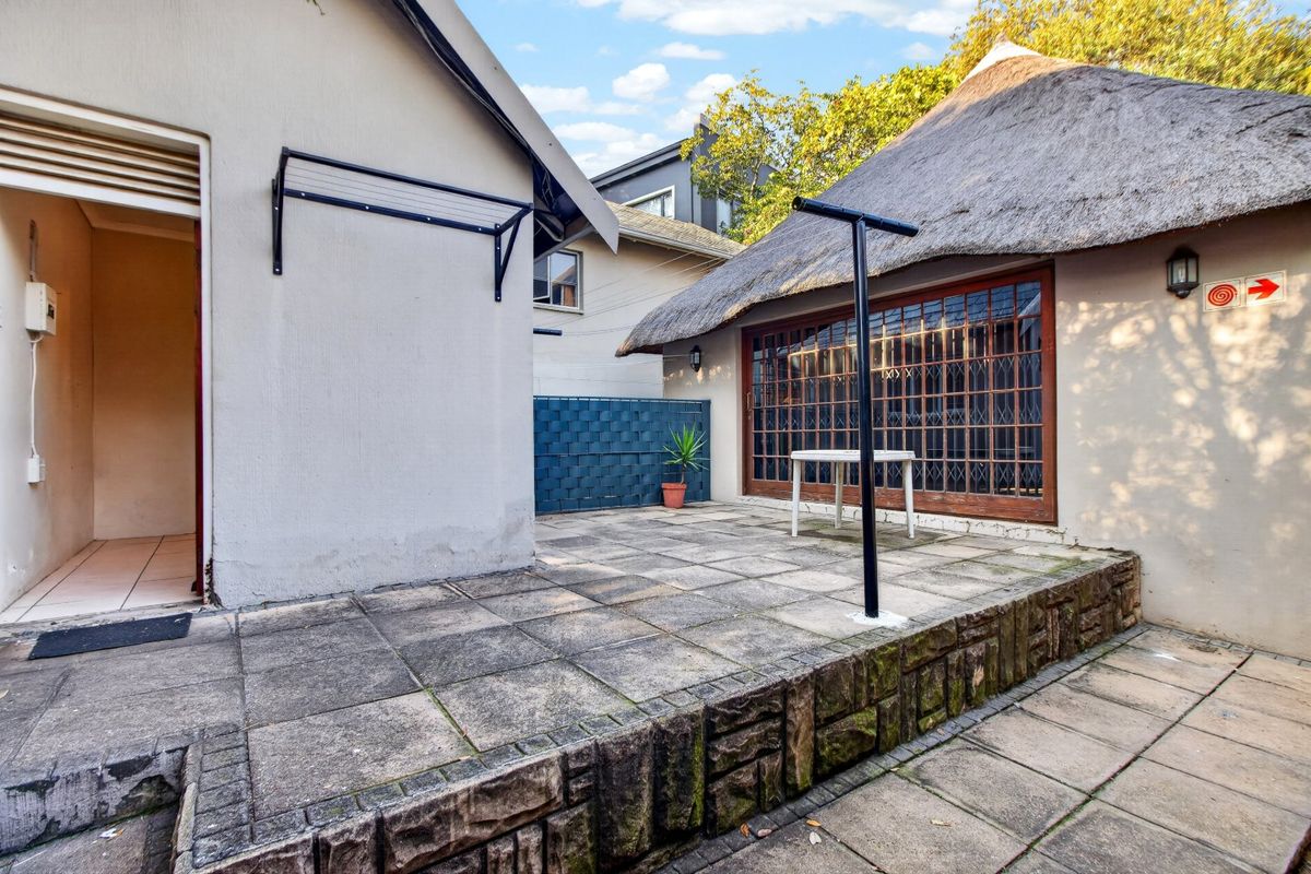 A paved rear patio area featuring a stone retaining wall, thatch-roofed structure with timber-framed doors, and practical wall-mounted and freestanding clotheslines.