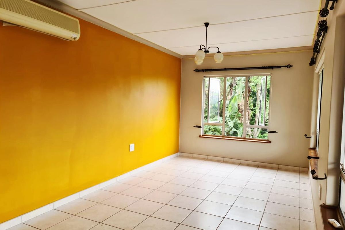 A bright, empty living room featuring tiled flooring, a split-system air conditioner, and large windows overlooking lush greenery.