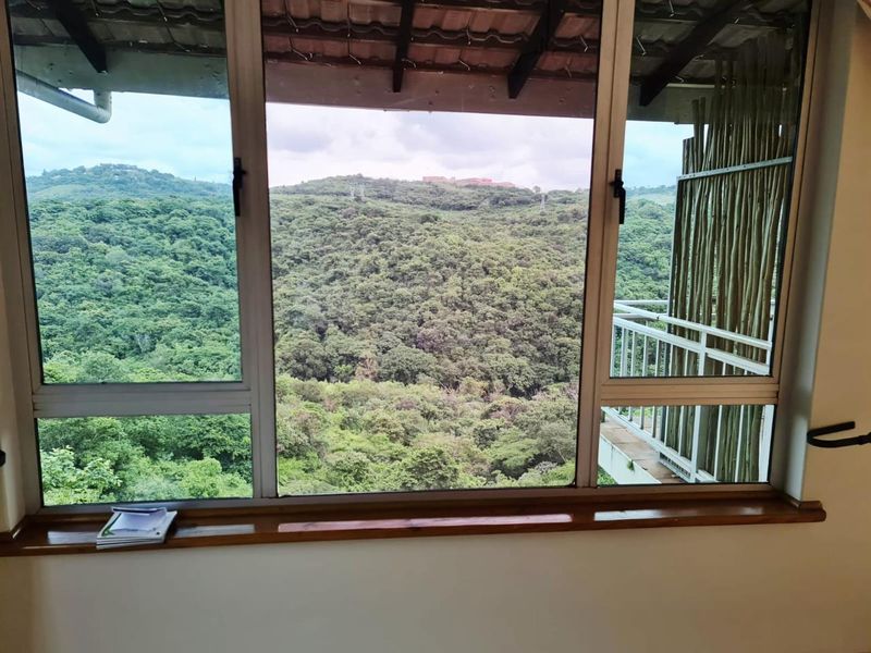 A view from an interior room looking out over a lush green forest landscape with a balcony visible to the side.