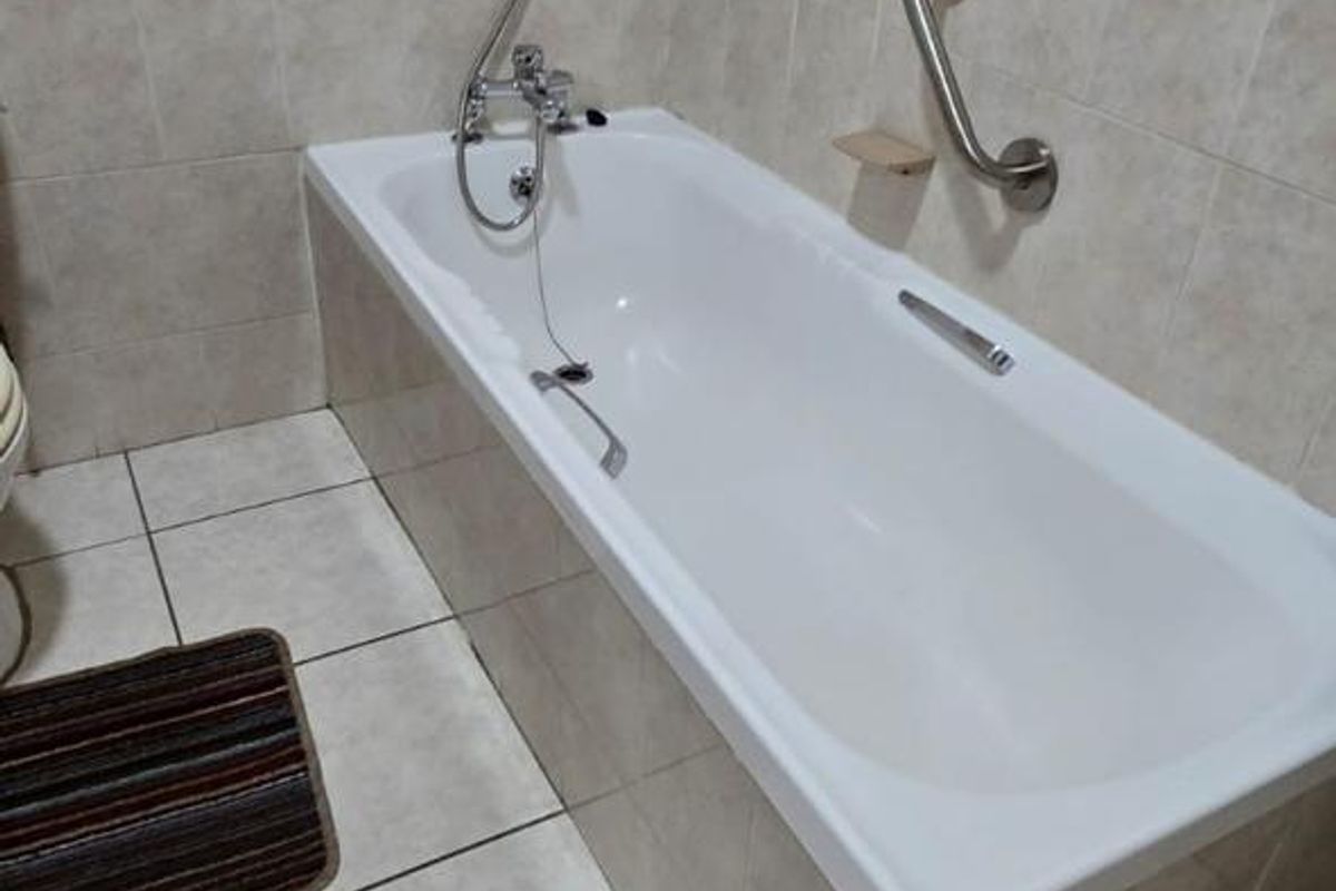 A functional bathroom featuring a built-in bathtub with a shower mixer and a wall-mounted grab rail, finished with neutral wall and floor tiles.