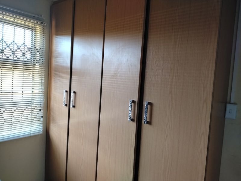 A bedroom featuring floor-to-ceiling built-in wooden cupboards, a window with venetian blinds, and security bars.