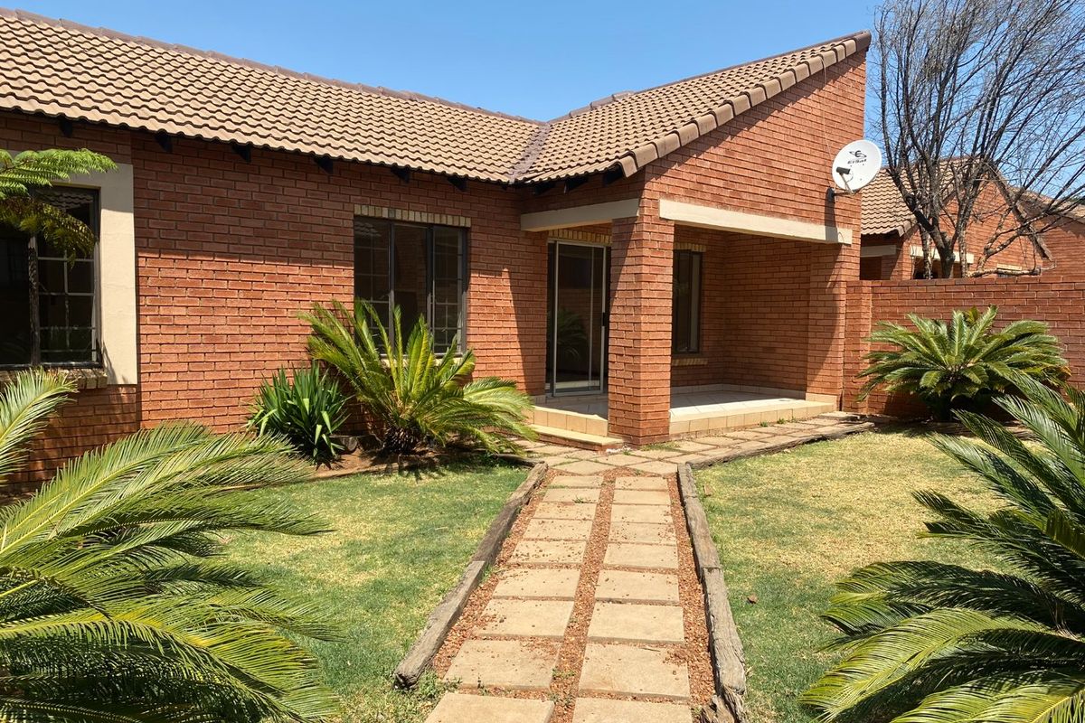 A well-maintained townhouse exterior featuring a classic brick facade, tiled roof, covered patio area, and a paved walkway leading through a landscaped garden.