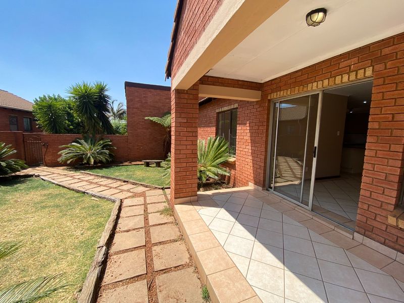 A covered brick patio featuring tiled flooring and a sliding glass door, overlooking a landscaped garden with a paved walkway.