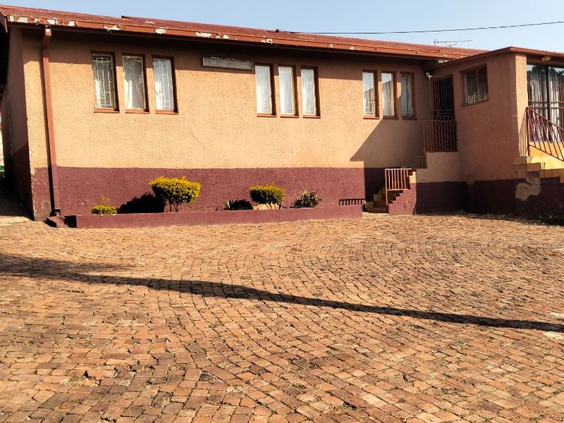 Front exterior view of a single-story house featuring a brick-paved driveway, security bars on windows, and visible wear to the exterior plaster and roof fascia.
