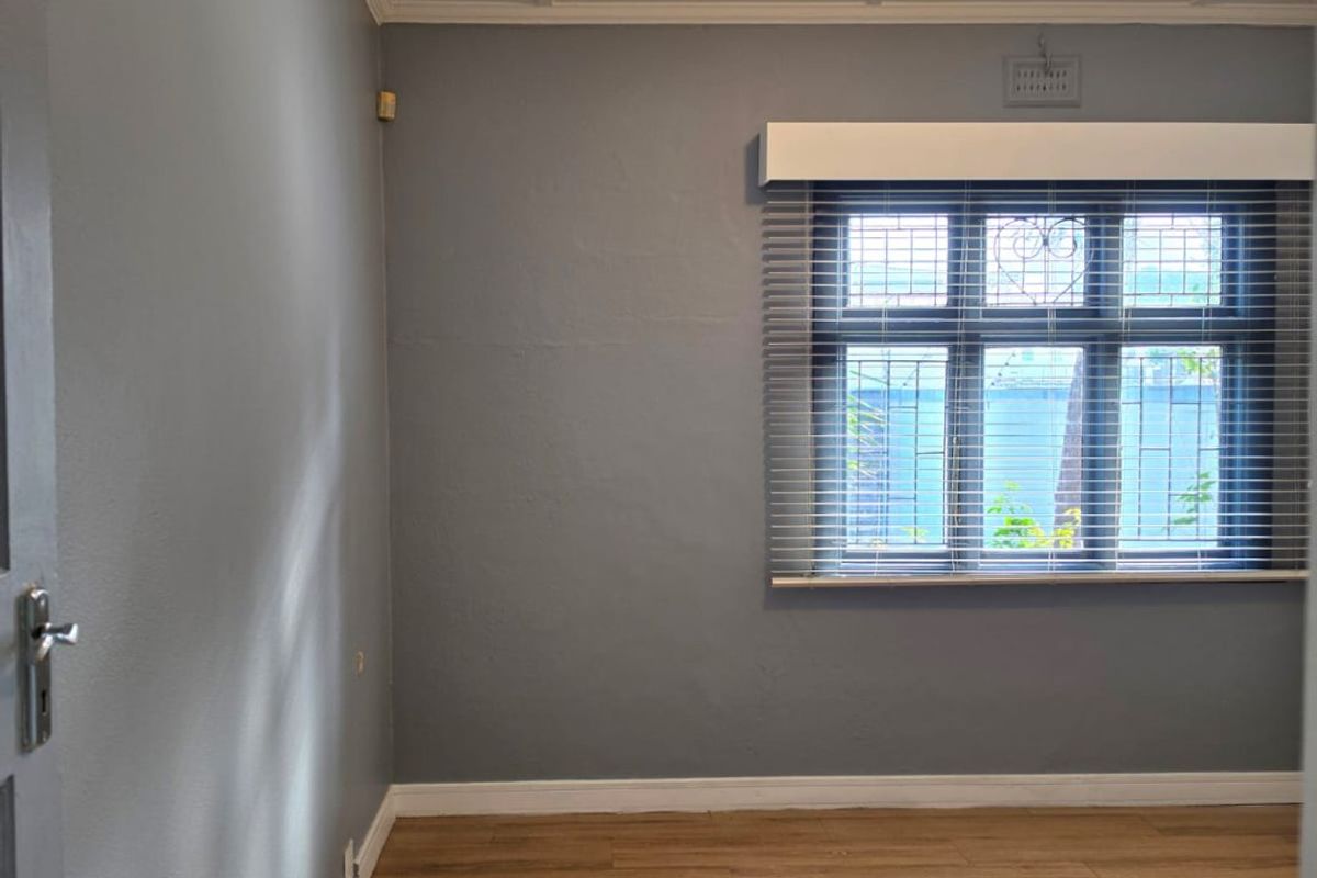 A bright, empty bedroom featuring modern grey walls, wood-look laminate flooring, and a window fitted with security bars and venetian blinds.