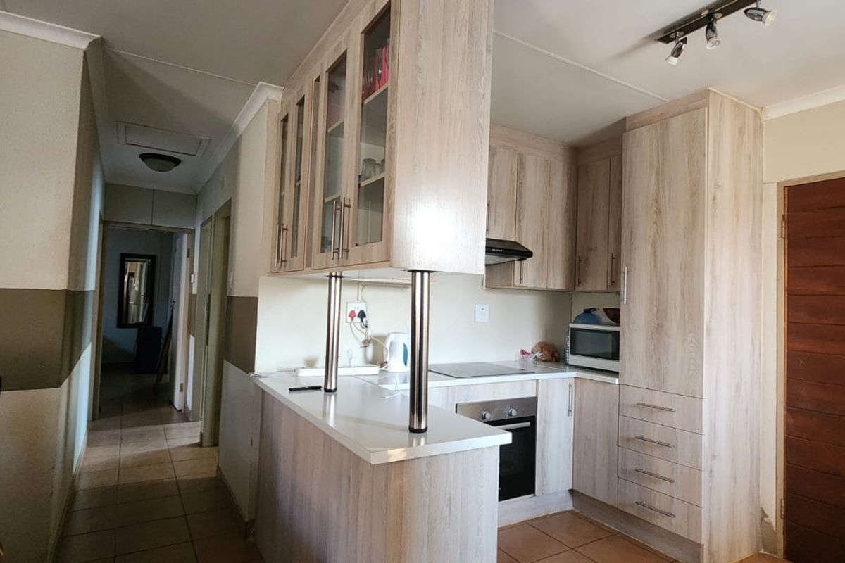 Compact kitchen featuring light wood-look cabinetry, integrated oven and hob, and a breakfast bar, leading into a tiled hallway.
